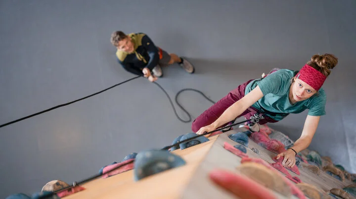 Eine Frau klettert in einer Kletterhalle, ihr Partner sichert sie. | © DAV / Julian Rohn